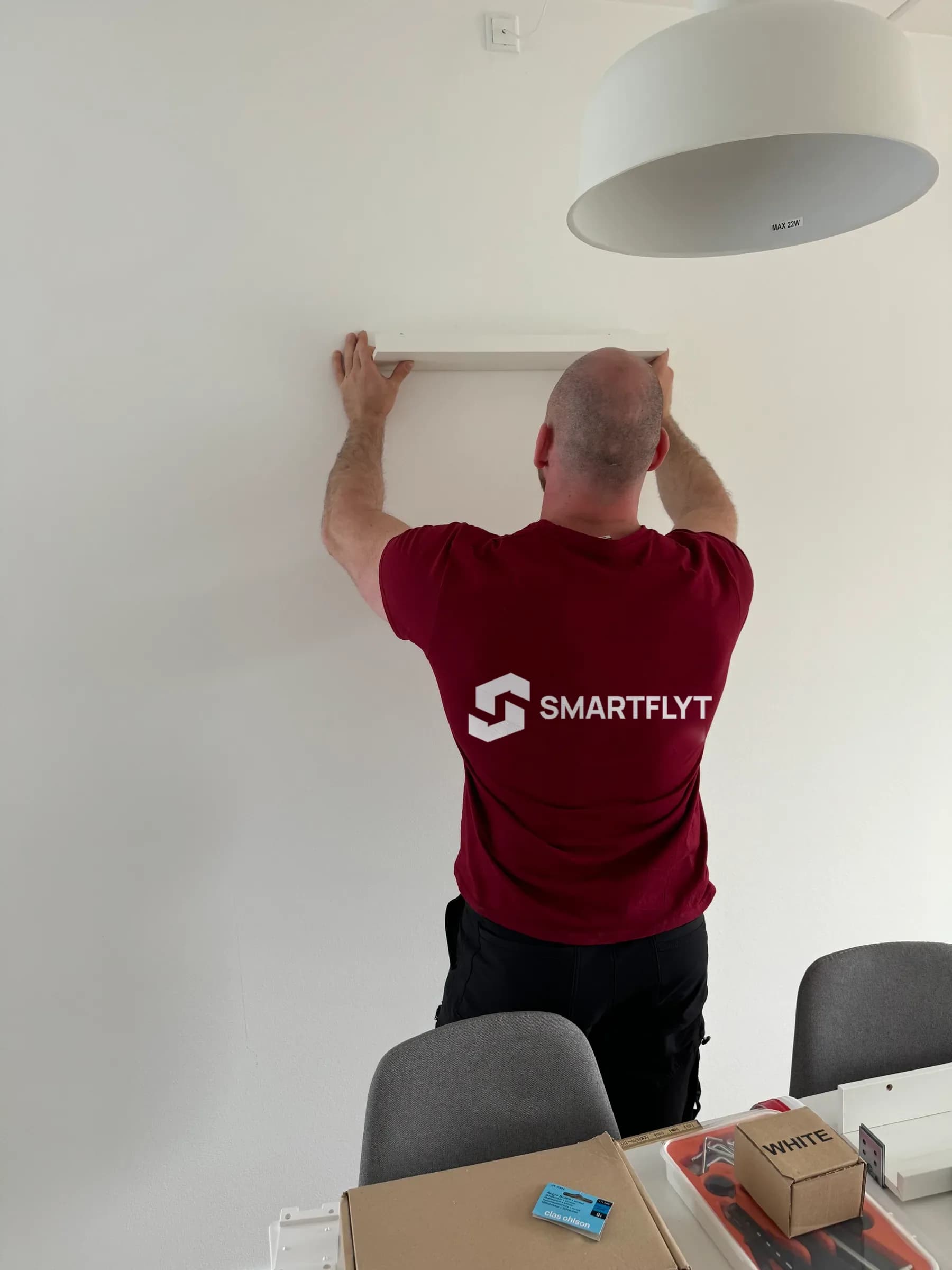 A mover in a Smart Flyt sweatshirt fixes a white shelf to the wall, with brackets and boxes on the table in front.