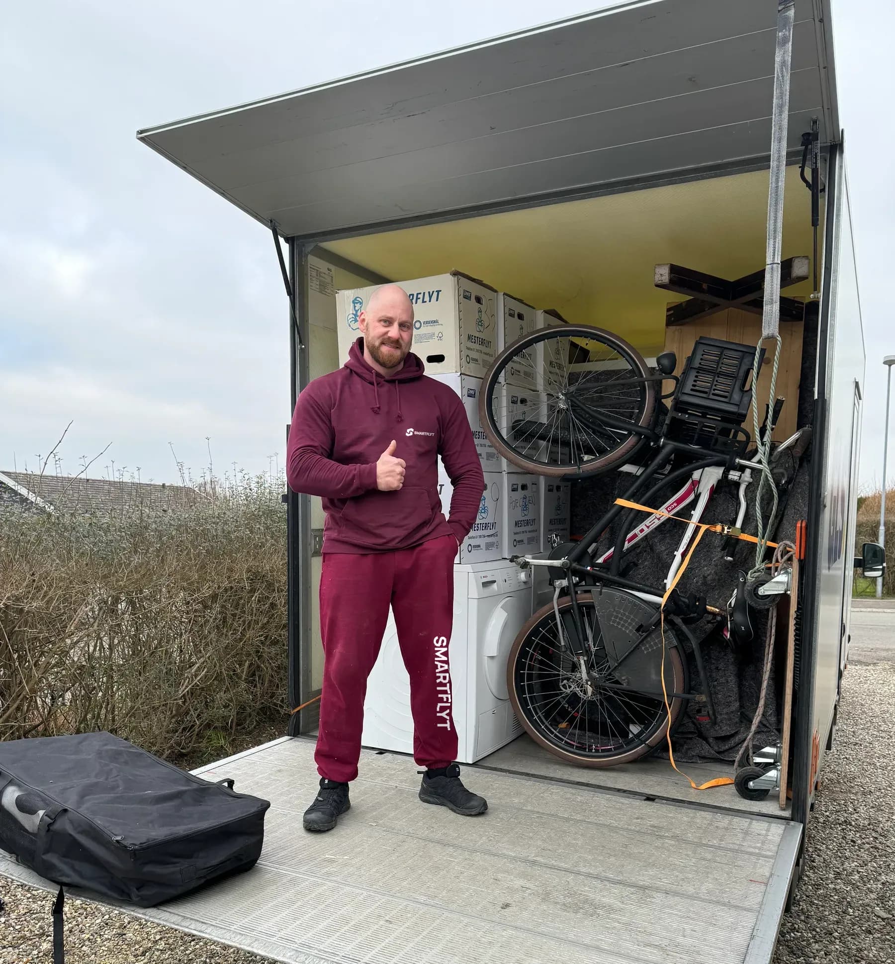 A mover in front of an open moving trailer filled with stacked moving boxes and wrapped furniture.