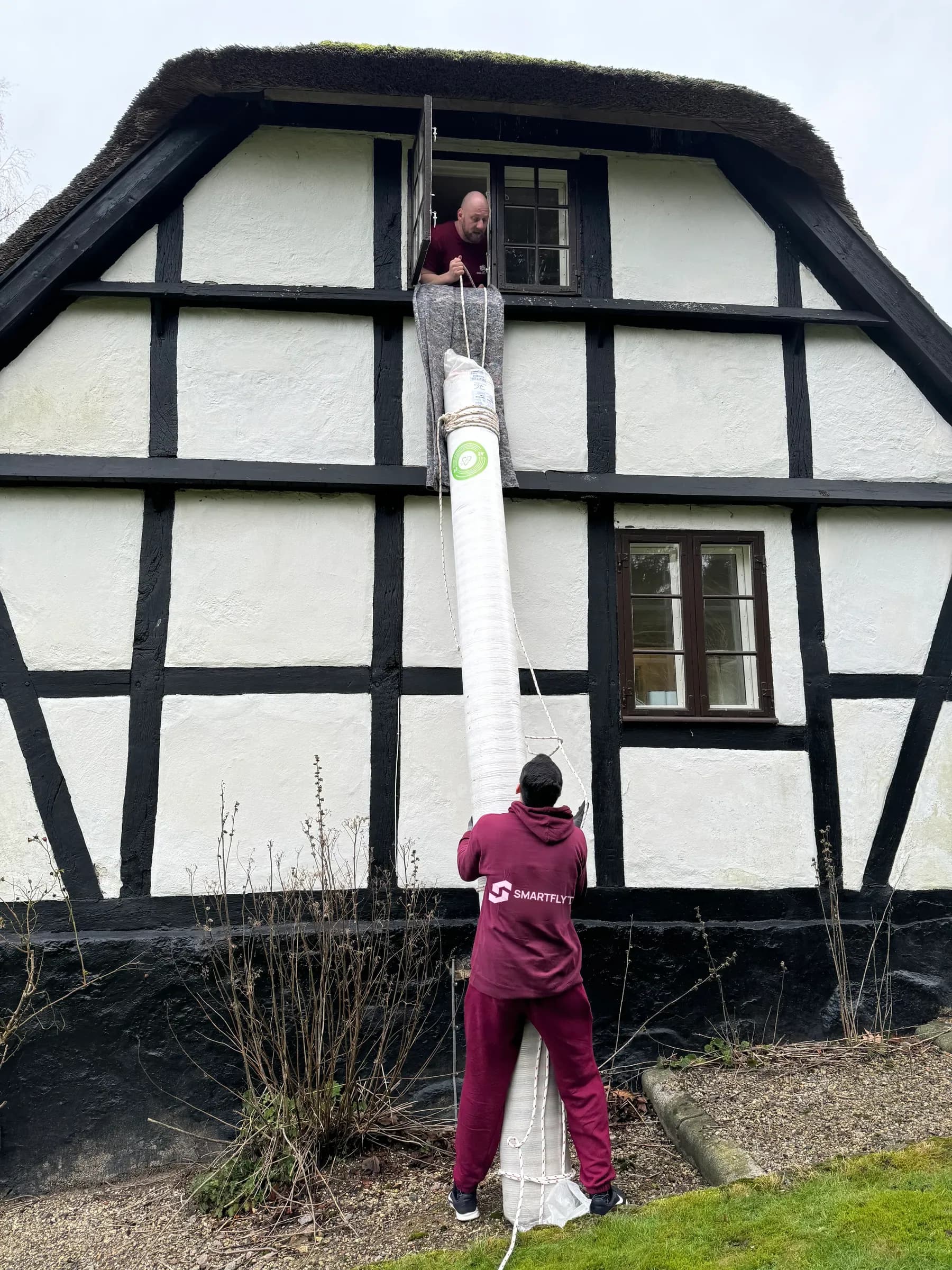 A wrapped piece of furniture is lowered by rope from a first floor window to a mover on the ground.